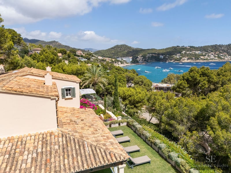 High-angle view of a villa with a tiled roof overlooking a garden, a turquoise bay with boats, and lush hills.