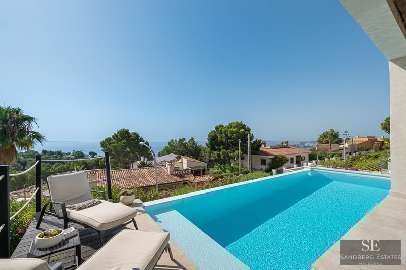 Infinity-Pool mit Sonnenliegen und Panoramablick auf mediterrane Häuser und das Meer unter blauem Himmel.