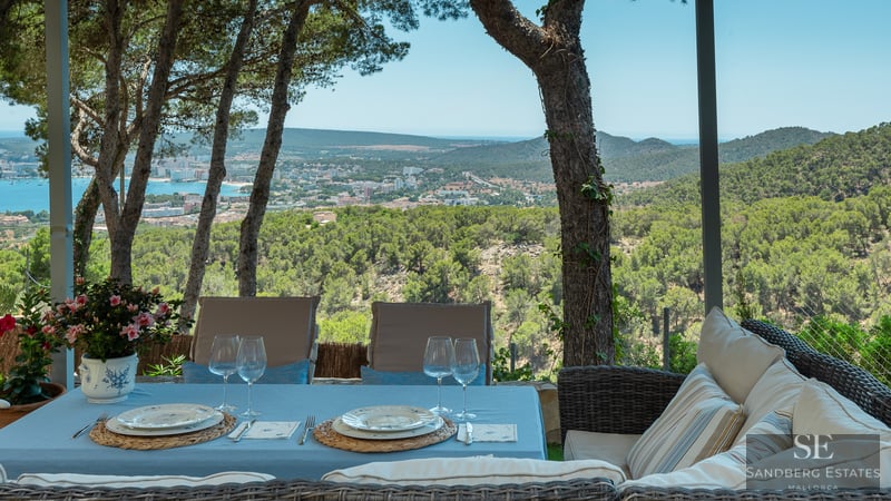 Matbord utomhus och korgsoffa på en skuggig terrass med utsikt över en kuststad och havet.