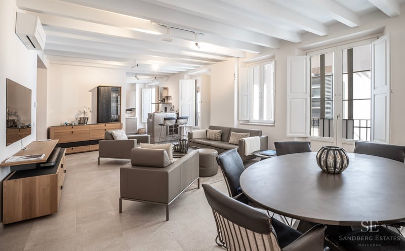 Elegant open-plan living room with gray sofas, white beamed ceiling, and natural light from large shuttered windows.