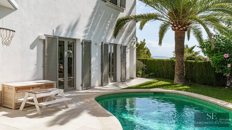Piscina turquesa junto a una villa blanca con una palmera, persianas grises y vistas lejanas al mar.
