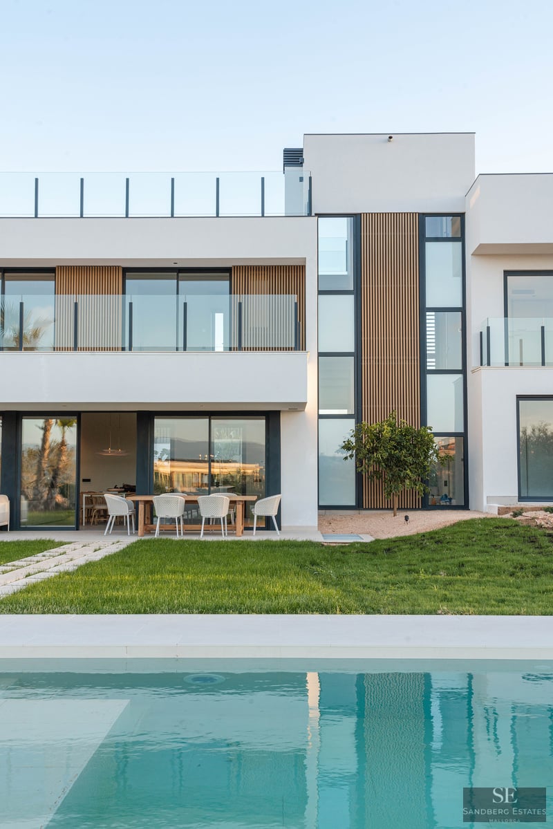 Modern white villa with wood accents, large windows, and a turquoise swimming pool in front of a green lawn.