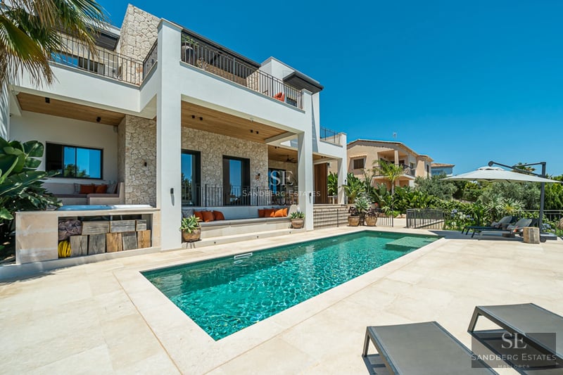 Vue extérieure d'une villa moderne avec piscine, pelouse soignée et grandes fenêtres réfléchissant la lumière du soleil.