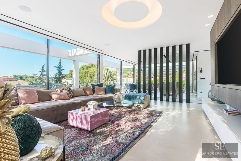 Modern living room featuring a large taupe sofa, colorful patterned rug, and floor-to-ceiling windows with greenery views.