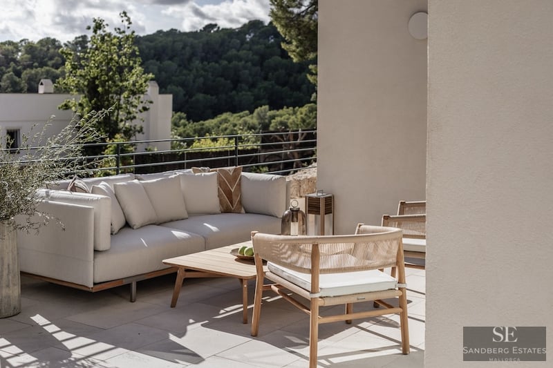 Modern outdoor seating area on a terrace with a white sofa, wooden chairs, and views of a lush green forest.