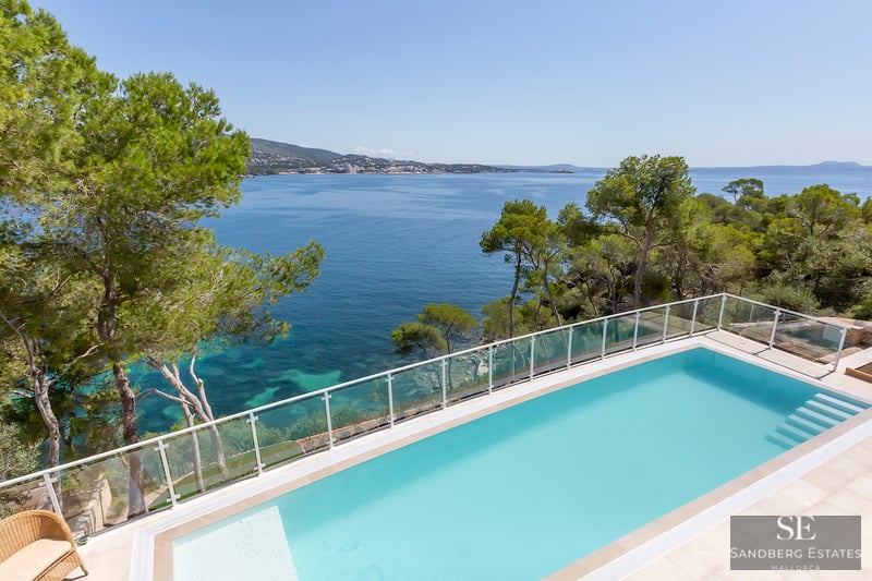 Rechteckiger türkisfarbener Pool mit Blick auf das Mittelmeer, umgeben von Kiefern und Glasgeländer.