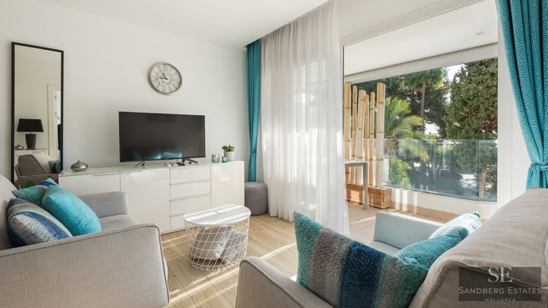 Modernt vardagsrum med grå soffor, turkosa kuddar och stora glasdörrar som leder ut till en lummig terrass.