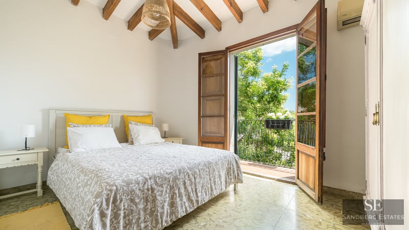 Ein helles Schlafzimmer mit sichtbaren Holzbalken und Fenstertüren, die auf einen sonnigen Balkon mit Bäumen führen.