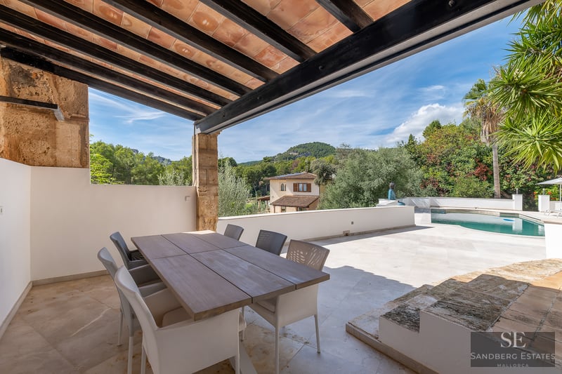 Outdoor dining area under a wooden beam pergola overlooking a pool, garden, and distant hills.