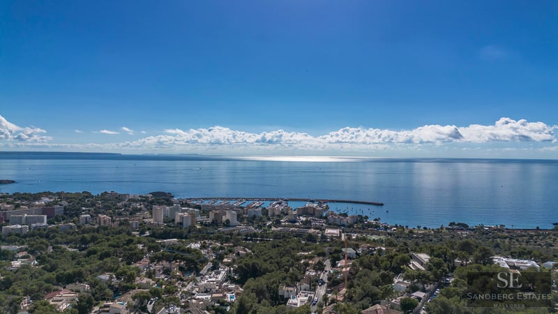 Vue aérienne d'une ville côtière avec marina, arbres verts et mer Méditerranée sous un ciel bleu clair.