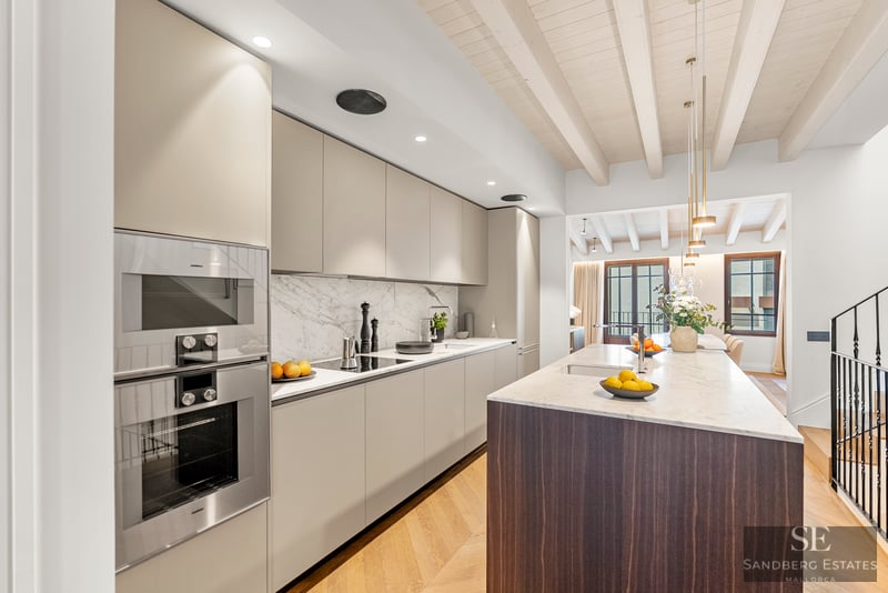 Cocina moderna con gabinetes beige, encimera de mármol blanco, isla de madera oscura y techo de vigas de madera clara.