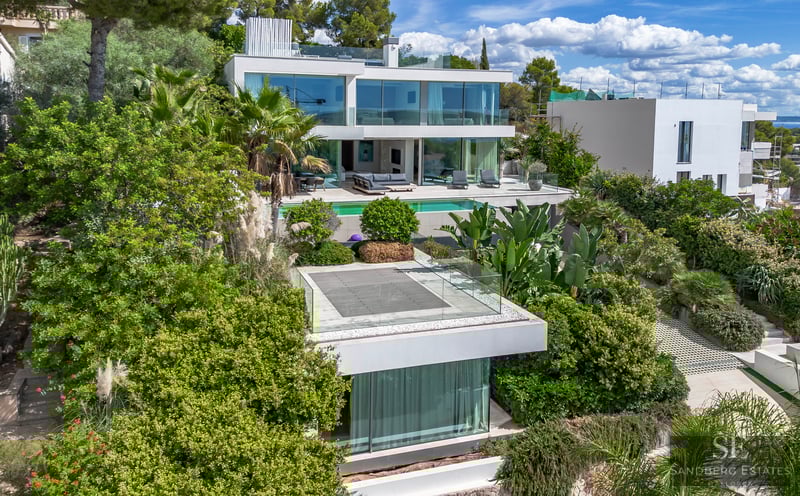 Modern multi-level white luxury villa with expansive glass walls, a turquoise pool, and dense Mediterranean landscaping.