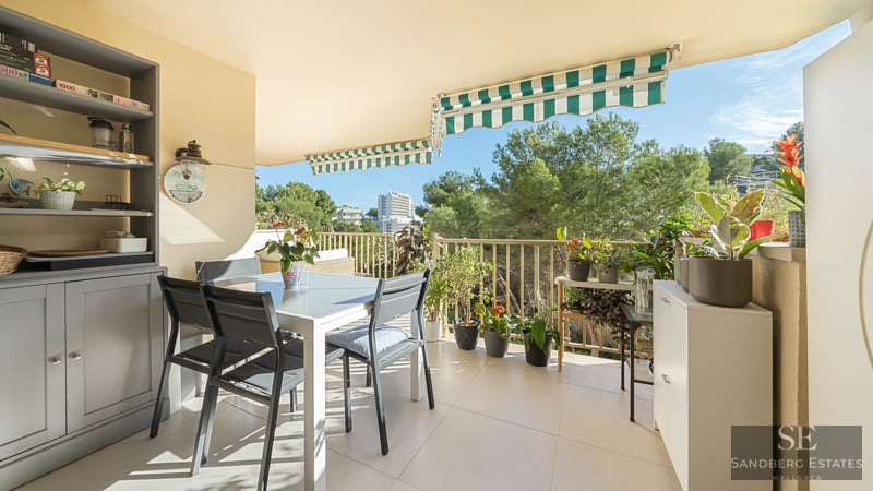 Un balcon ensoleillé avec une table à manger blanche, des chaises sombres, de nombreuses plantes en pot et des stores rayés.
