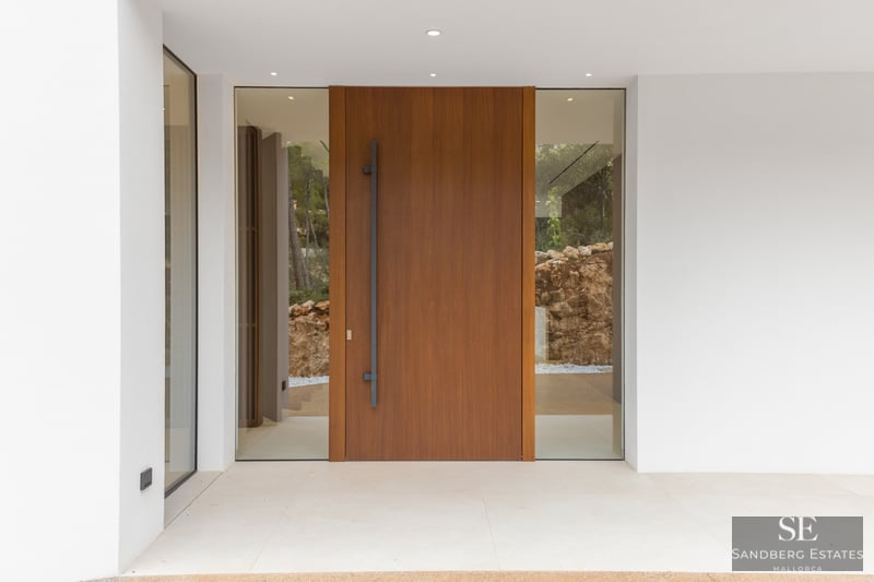 Large wooden pivot door with glass side panels flanked by smooth white walls and minimalist lighting.