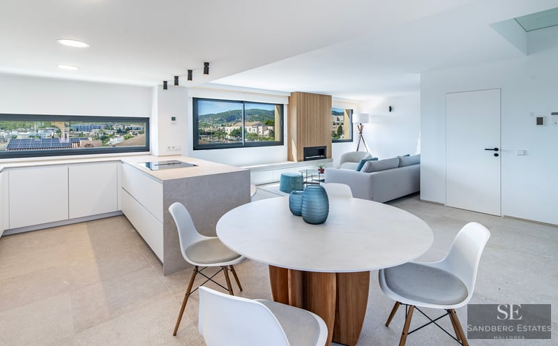 Espacio abierto luminoso con comedor, isla de cocina moderna y salón con grandes ventanales y vistas al pueblo.