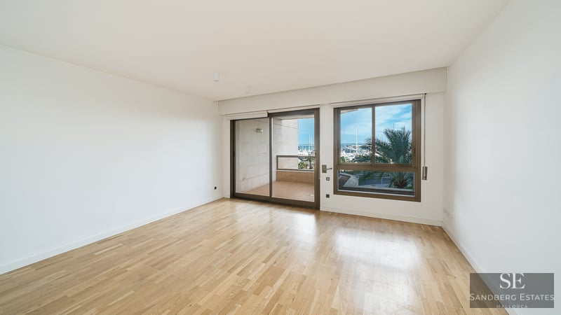 Empty modern room with light wood floors and large windows overlooking a balcony and a marina with palm trees.