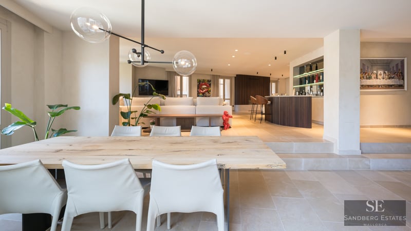 A spacious modern open-plan room featuring a wooden dining table, white chairs, glass globe pendants, and a bar area.