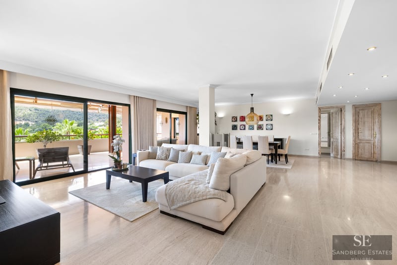 Salon spacieux avec canapé blanc en L, sol en marbre et grandes baies vitrées ouvrant sur une terrasse paysagée.