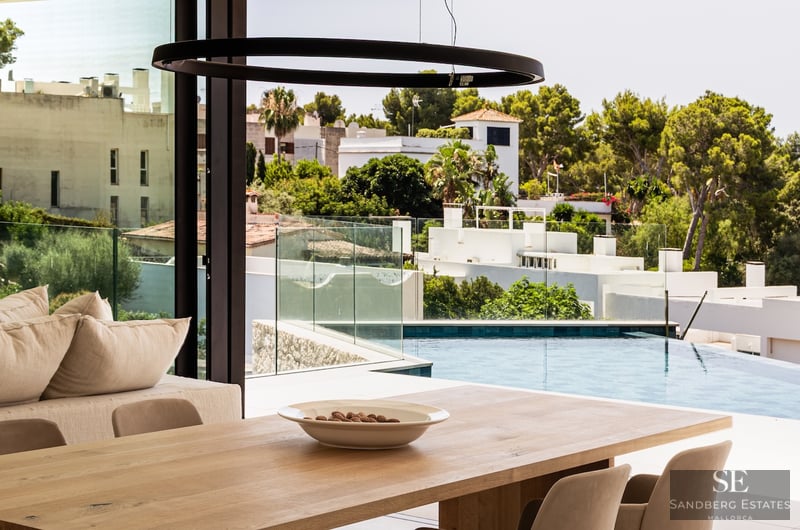 A minimalist dining room with a wooden table and glass walls overlooking a bright blue infinity pool and greenery.