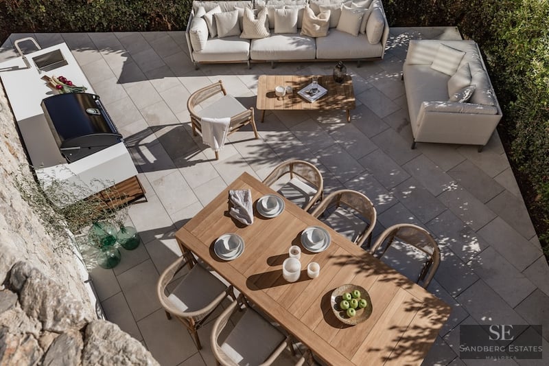 Overhead view of a modern stone patio featuring a teak dining set, white sofas, and a sleek outdoor kitchen with BBQ.