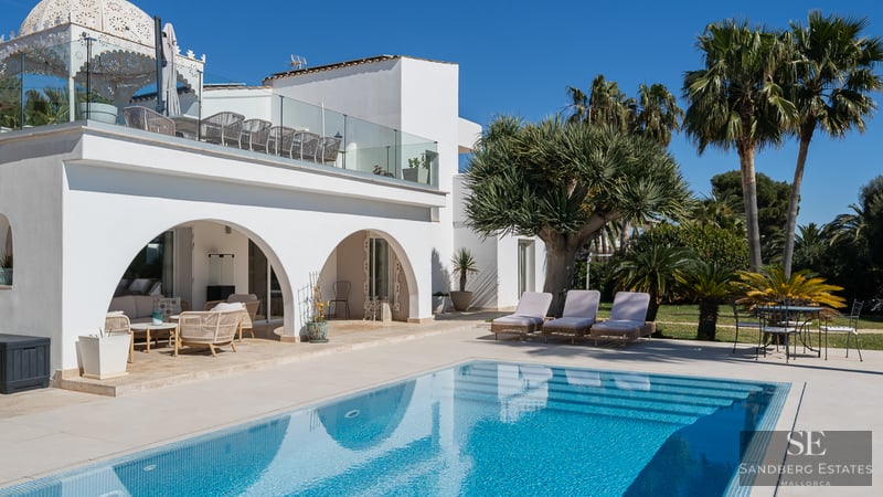 Villa blanca con arcos frente a una piscina turquesa con tumbonas y palmeras bajo un cielo azul despejado.