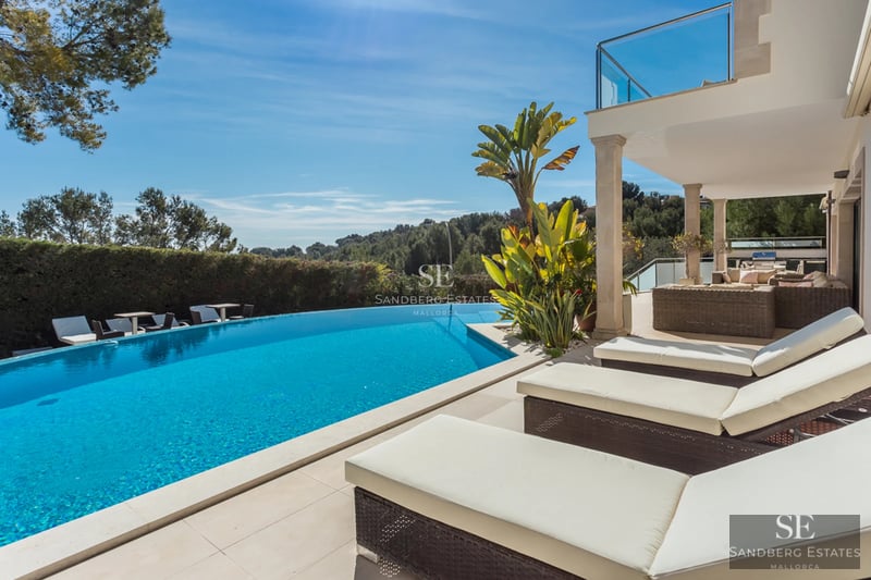 Exterior view of modern villa with infinity pool, lush garden, and minimalist architecture. White facade with large windows and natural light.