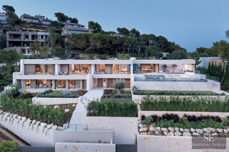 Villa moderna de dos plantas con piscina infinita, jardines de piedra y fachada de cristal iluminada al anochecer.