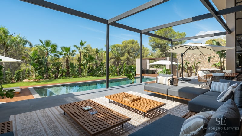 Modern outdoor lounge with grey sofas and wooden tables next to a swimming pool and lush tropical garden.
