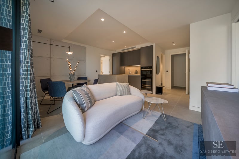 Salón moderno con sofá curvo blanco, alfombra azul y mesa de comedor, junto a una cocina gris oscuro.