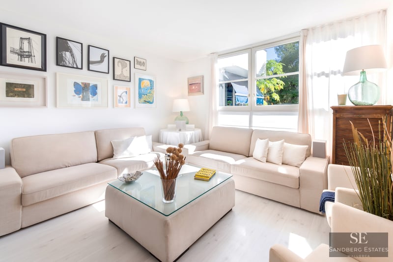 Helles Wohnzimmer mit zwei beigen Sofas, einem Glastisch, großen Fenstern und einer Wand mit gerahmter Kunst.