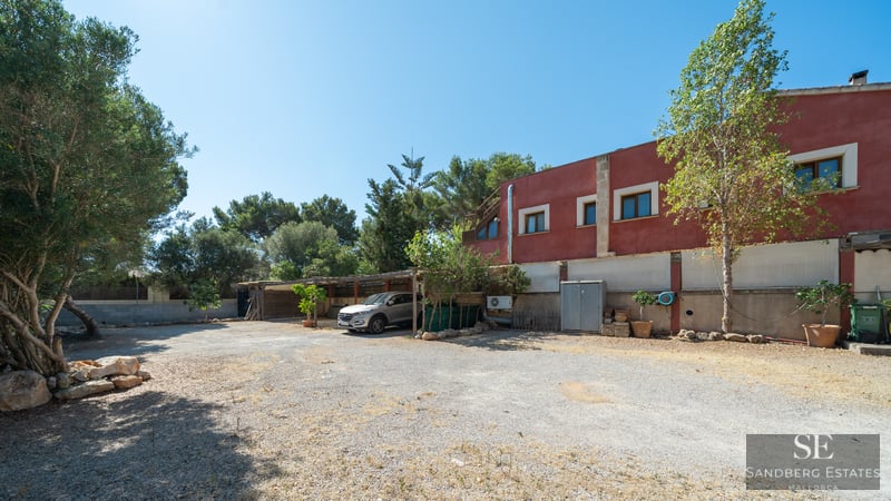 A wide gravel parking area in front of a red building with a silver car parked under a wooden carport.