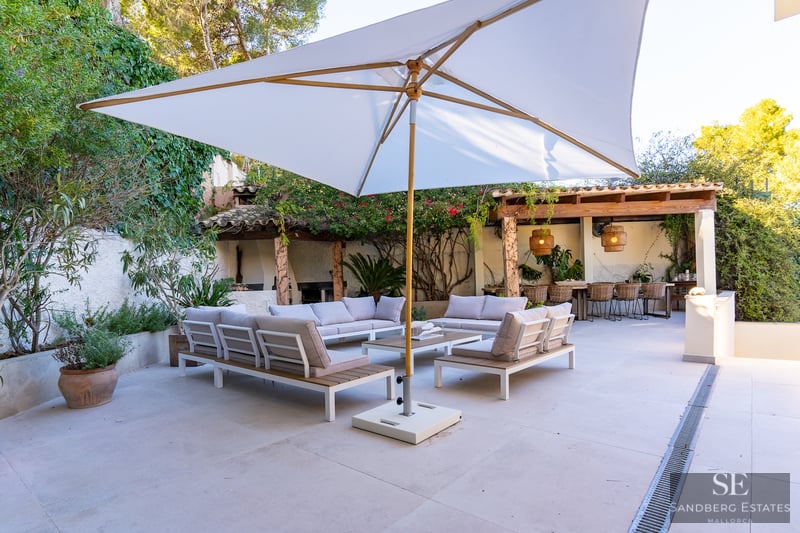 Spacious terrace featuring modern sofas, a large white parasol, and a dining area under a pergola surrounded by greenery.