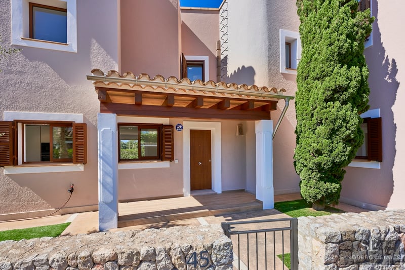 Entrance of a beige villa with a wooden door, stone wall, and a tall cypress tree under a bright blue sky.