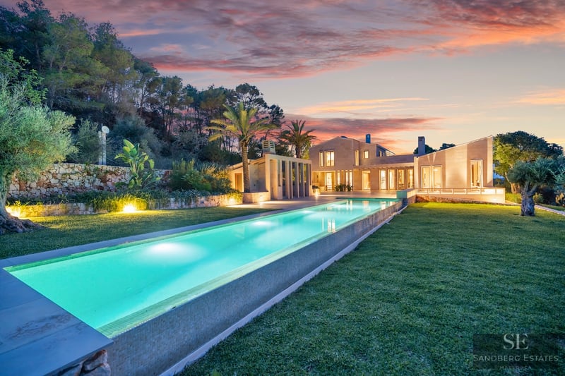 Long glowing lap pool leading to a modern stone villa under a vibrant orange and pink sunset sky.