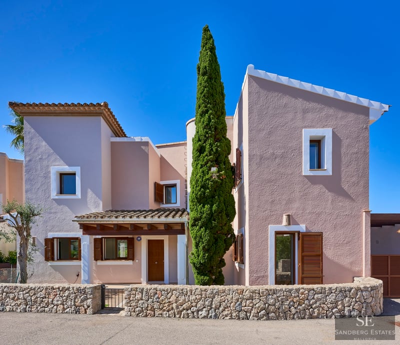 Front view of a pink two-story Mediterranean villa with wooden shutters, a stone wall, and a tall cypress tree.