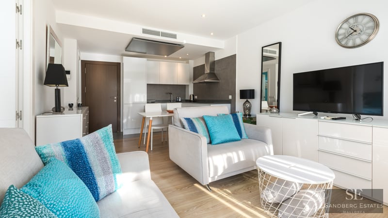 Ljust vardagsrum med grå fåtölj, turkosa kuddar, TV-bänk och modernt kök i bakgrunden.