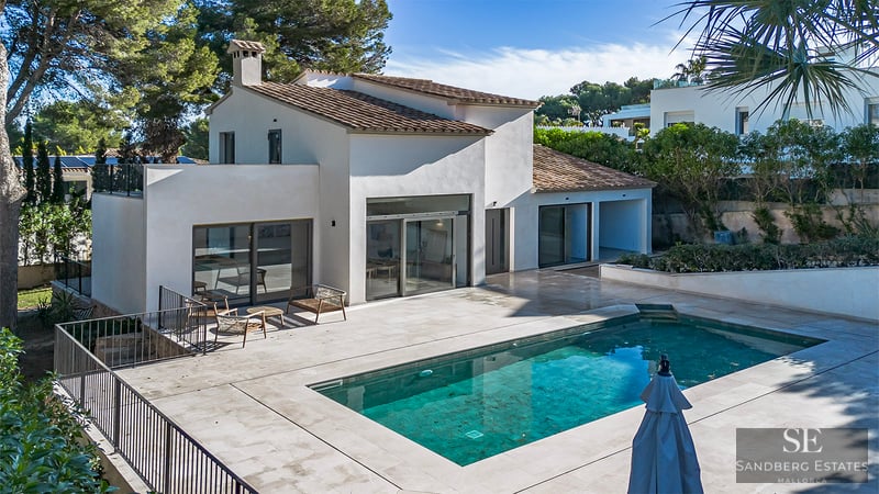 White Mediterranean-style villa with a rectangular blue swimming pool and a large stone deck under bright sunlight.