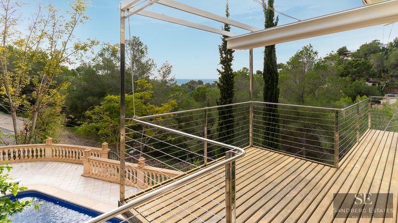 Plataforma de madera elevada con barandillas metálicas con vistas a una piscina y bosque verde bajo el cielo azul.