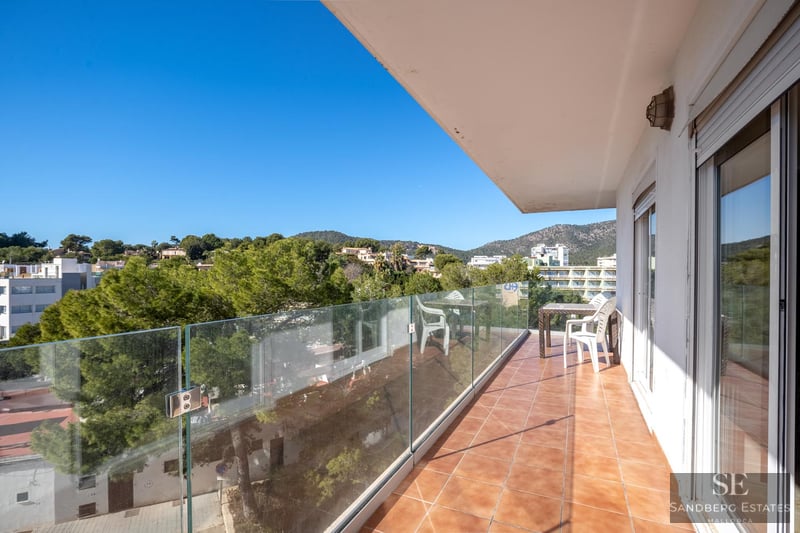 A long balcony with glass railings and terracotta tile flooring overlooking lush green hills under a clear blue sky.