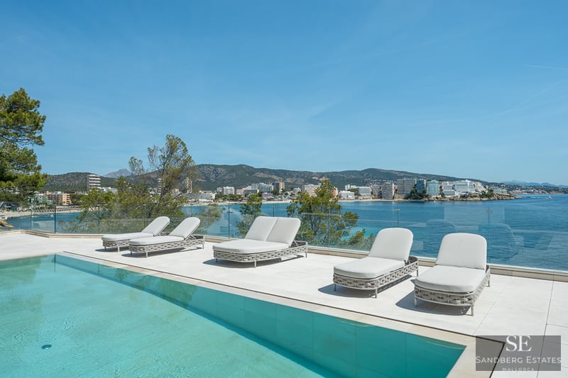 Modern white terrace with a turquoise pool, loungers, and a glass railing overlooking a scenic Mediterranean bay.