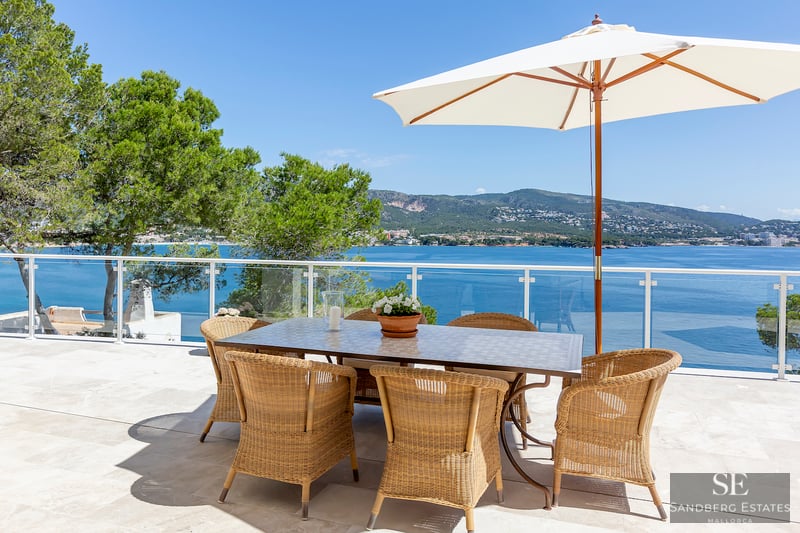 Außen-Essbereich mit Korbstühlen und weißem Sonnenschirm auf Steinterrasse mit Blick auf das blaue Meer und Berge.