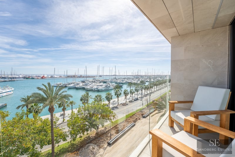 Modern balcony with wooden chairs and glass railing overlooking a marina full of yachts and palm trees.
