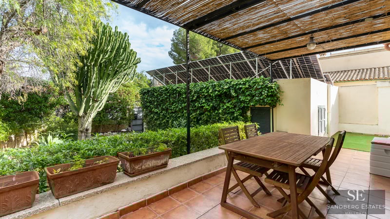 Wooden dining table under a bamboo pergola on a terracotta terrace surrounded by lush greenery and solar panels.