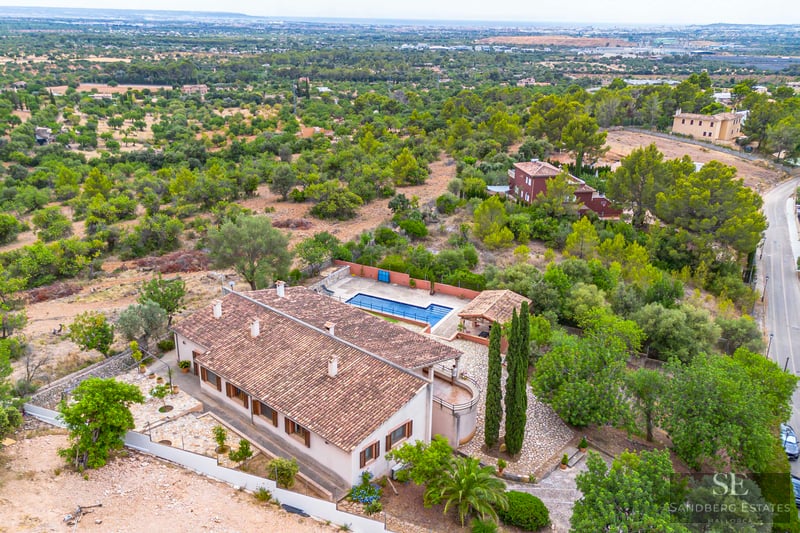 Vista de pájaro de una villa con tejado de terracota, piscina azul y amplios jardines verdes.