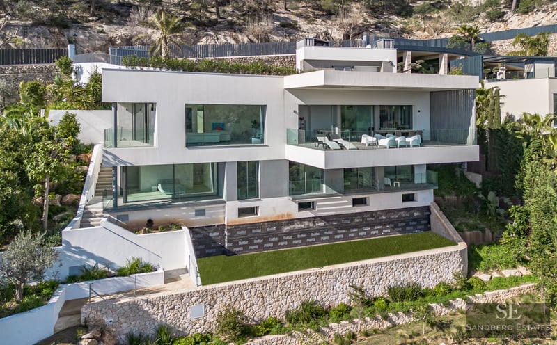 Modern white multi-story villa with large glass windows and terraces built into a rocky hillside with stone walls.