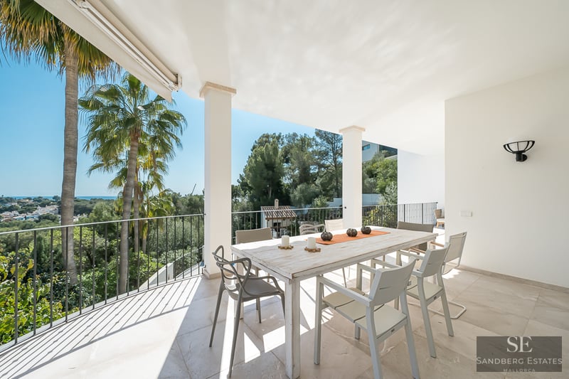 Terrasse blanche avec table à manger en bois, chaises modernes et vue sur les palmiers et la mer.