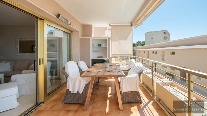 A sunlit balcony terrace with a wooden dining table, wicker chairs, and a metal railing overlooking urban buildings.