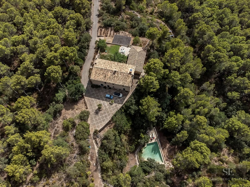 Aerial view of a stone house with terracotta roof, courtyard, and a pool, all surrounded by a dense green pine forest.