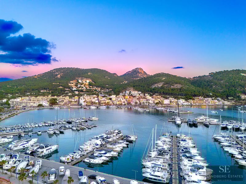 Aerial view of a luxury marina filled with white yachts under a pink and blue sky next to green mountains.