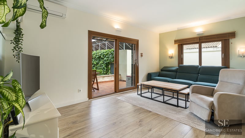 Bright living room with teal sofa, armchair, wood floors, and glass doors leading to a lush green terrace.